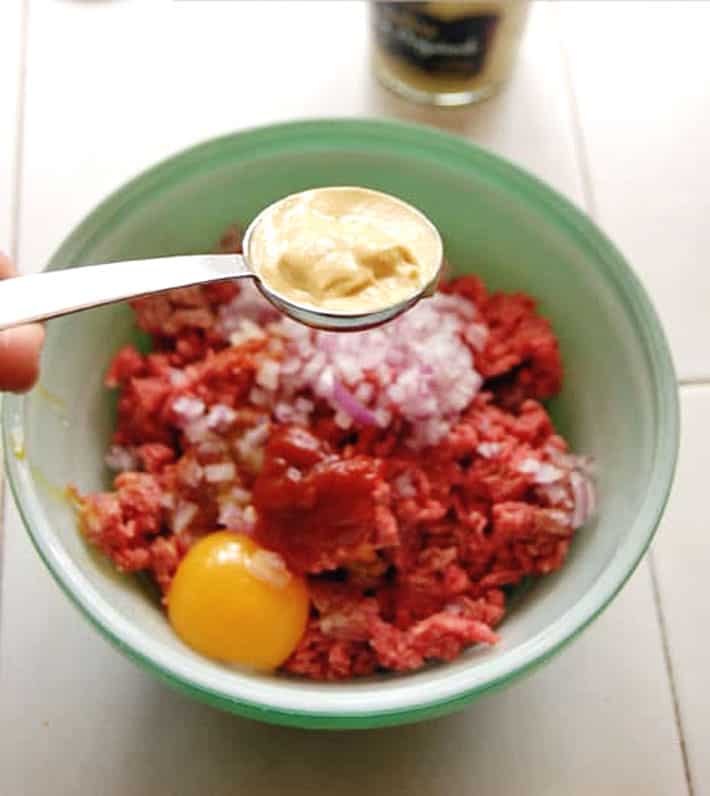 Dijon mustard in metal tablespoon hovering over green bowl with ground beef and other hamburger ingredients. 