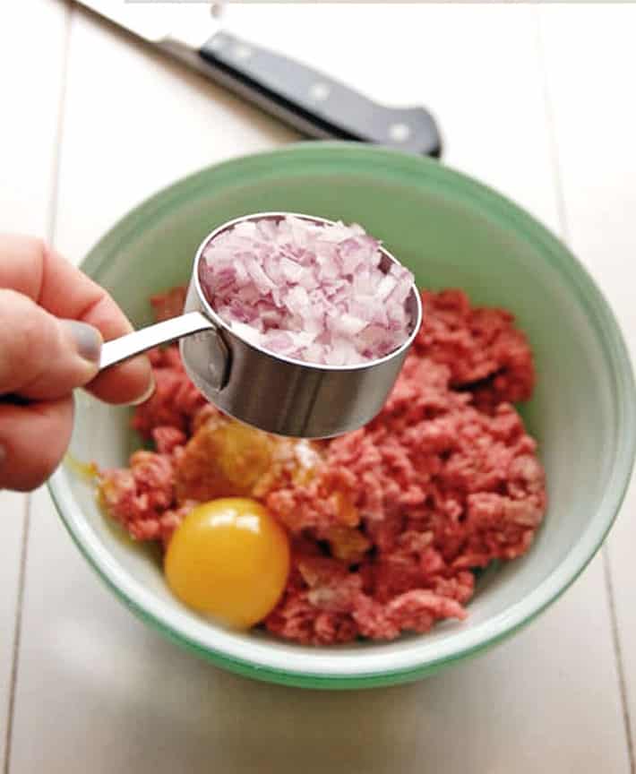 Finely diced onions in metal measuring cup over bowl with ground beef.