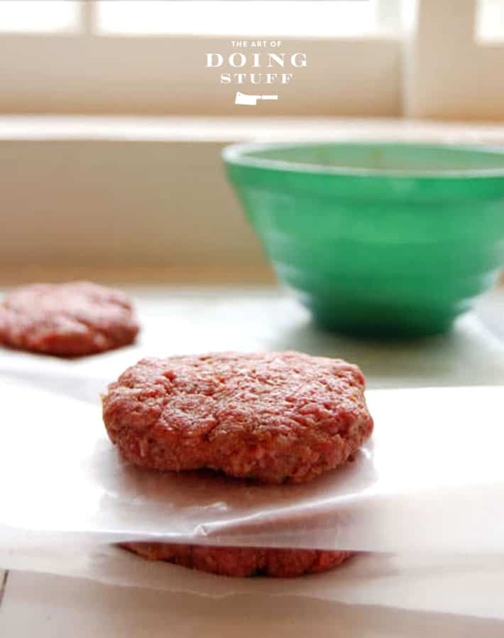 Hand formed hamburger patties in between layers of waxed paper on white tile countertop.