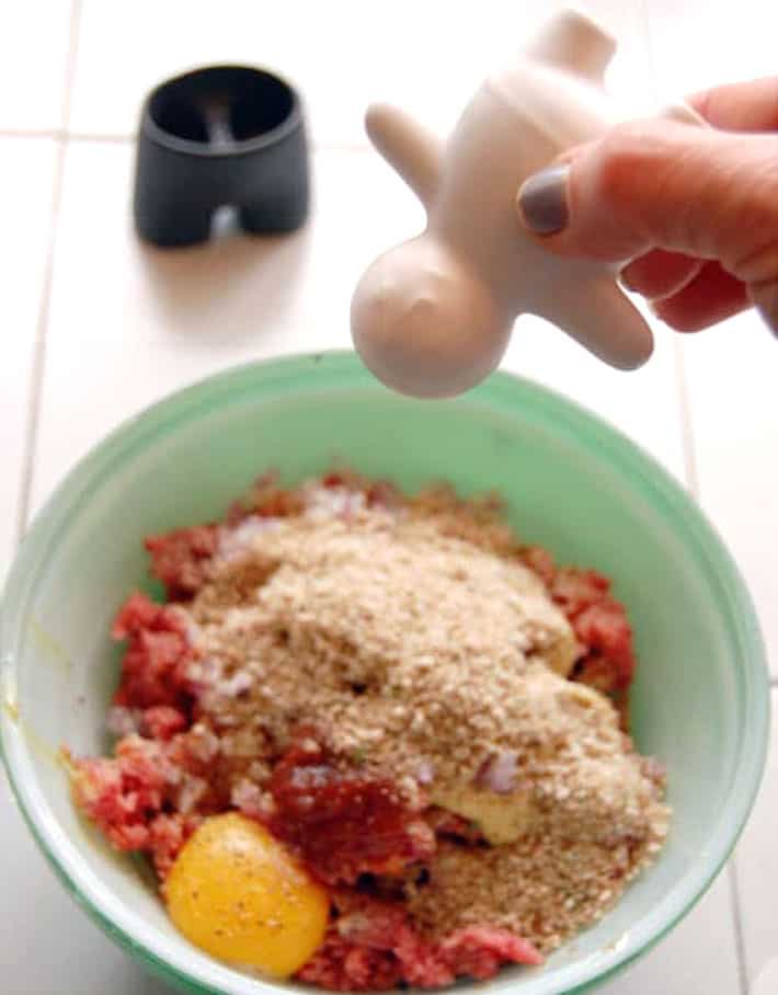 Shaking man shaped salt shaker into green bowl with unmixed hamburger ingredients.