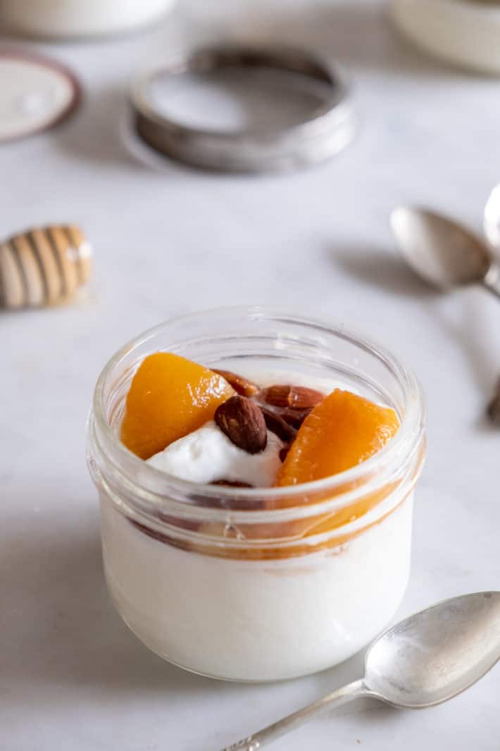 A short mason jar filled with homemade yogurt topped with home canned peaches and crunchy almonds.