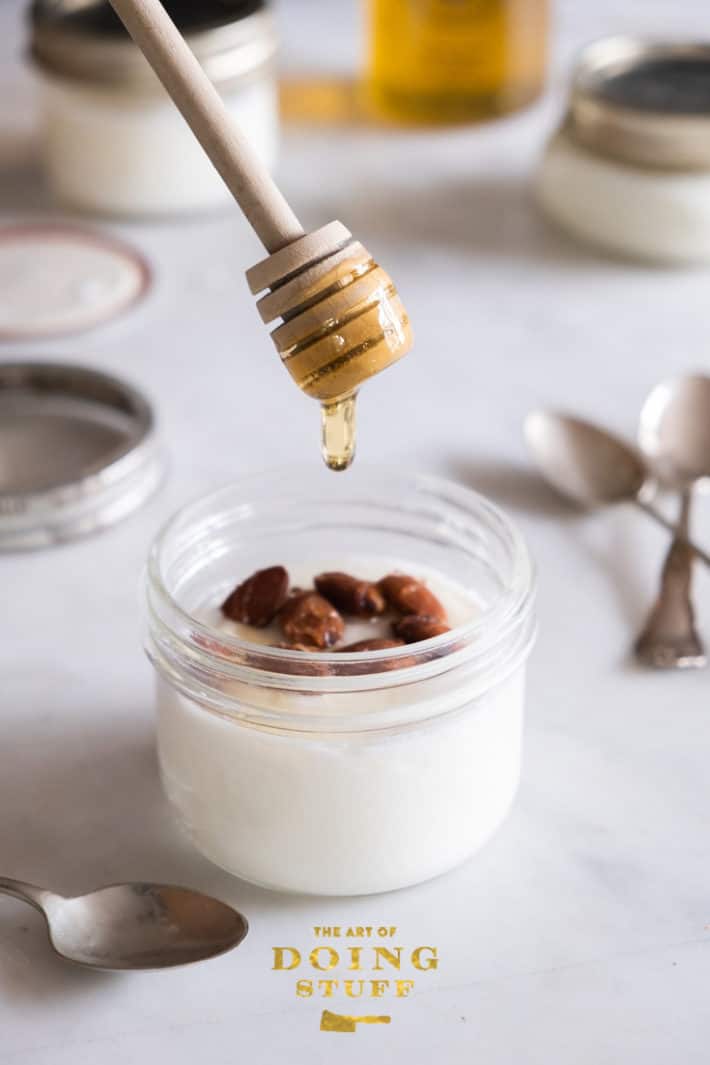 Golden honey dripping off a wood honey dripper into a small mason jar of yogurt topped with almonds.