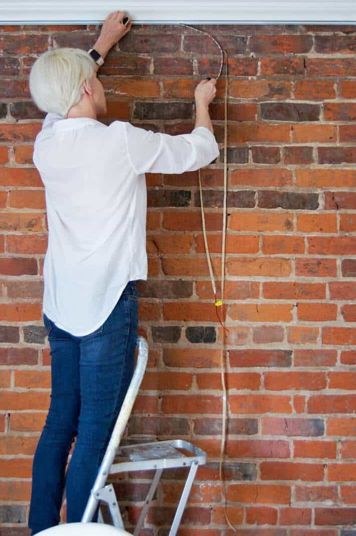 Woman installing strip lights under crown moulding to illuminate brick wall.