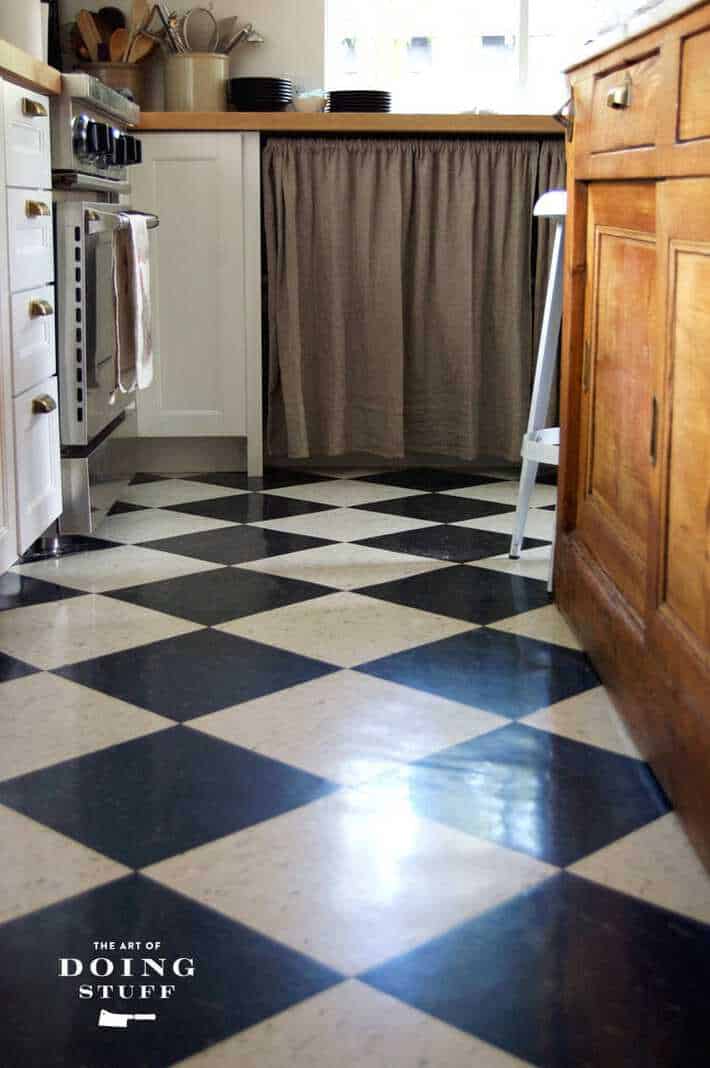 A modern country kitchen with a VCT tile black and white check floor, white shaker cabinets and a linen curtain under the counter..