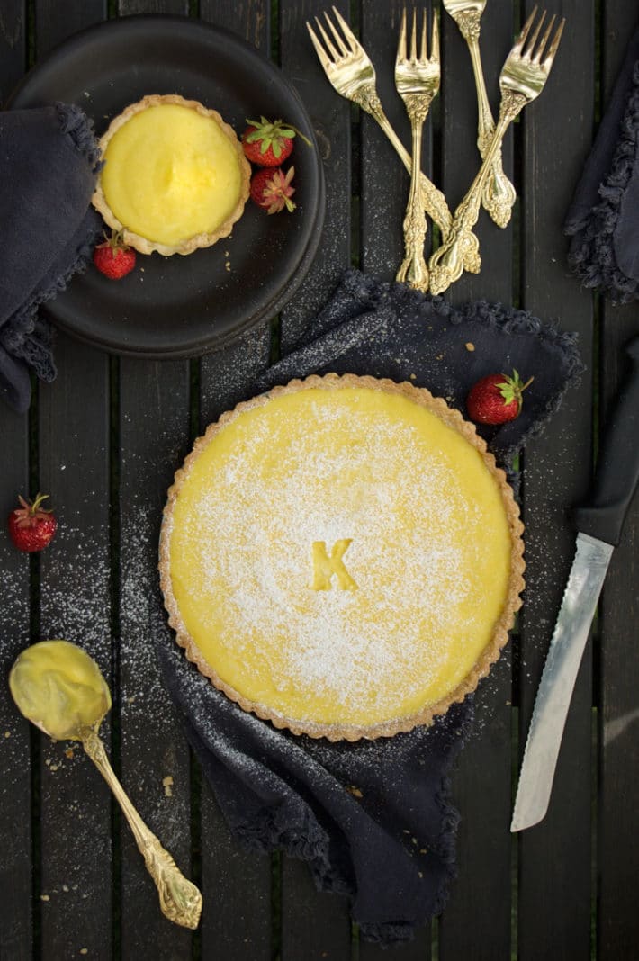 A lemon tart sprinkled with powdered sugar on a black table surrounded by gold flat wear, black napkins and bright red strawberries.