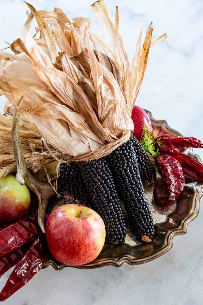 A fall harvest centerpiece with black corn, apples and dried peppers on a tarnished silver platter.