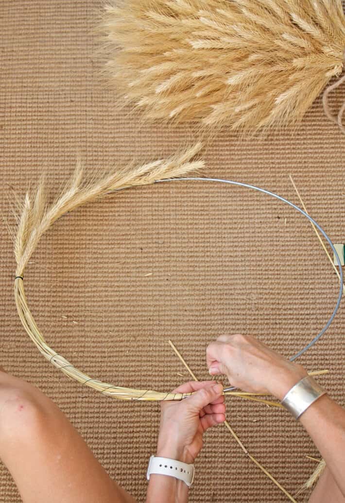 Half completed simple wheat wreath laying on natural rug with wheat sheaf in corner.
