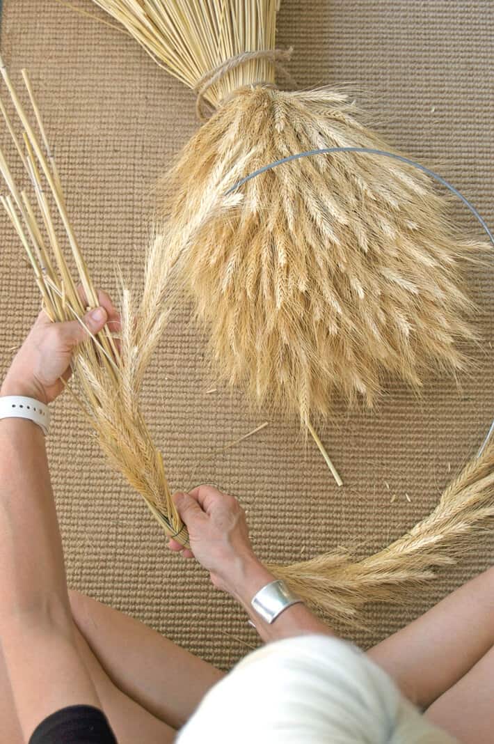 A woman grasps the round wire form of a wheat wreath, while weaving wheat into it.