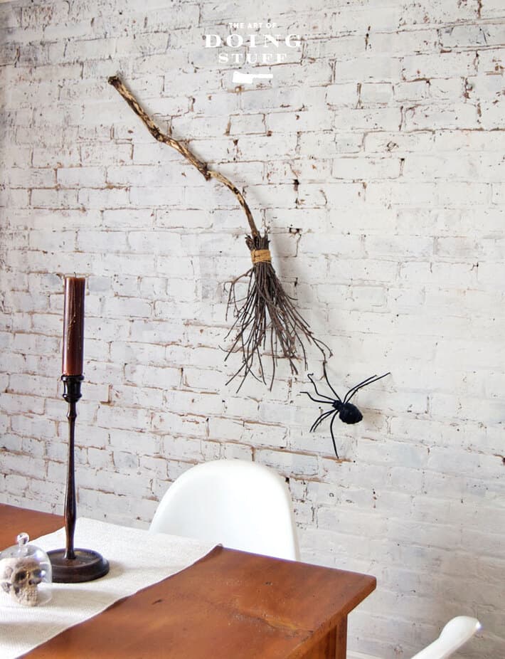 A witches broom appears suspected on a painted white brick wall with a large spider climbing towards it.
