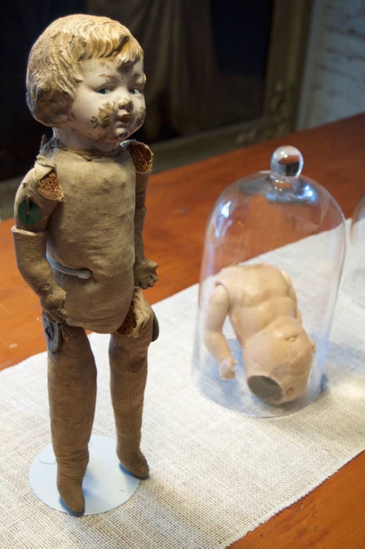 An antique straw-filled doll with porcelain head displayed next to a doll's torso under glass on a dining room table for Halloween.