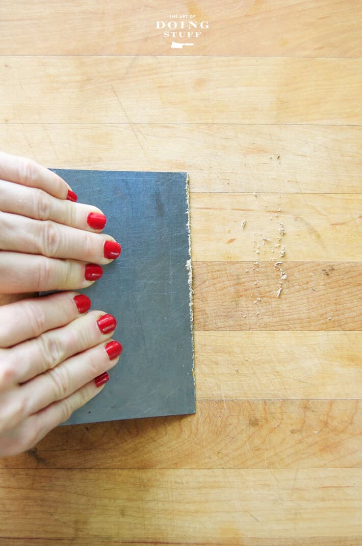 Woman's hands with red nailpolish run a dough scraper across a wood counter to remove guck.