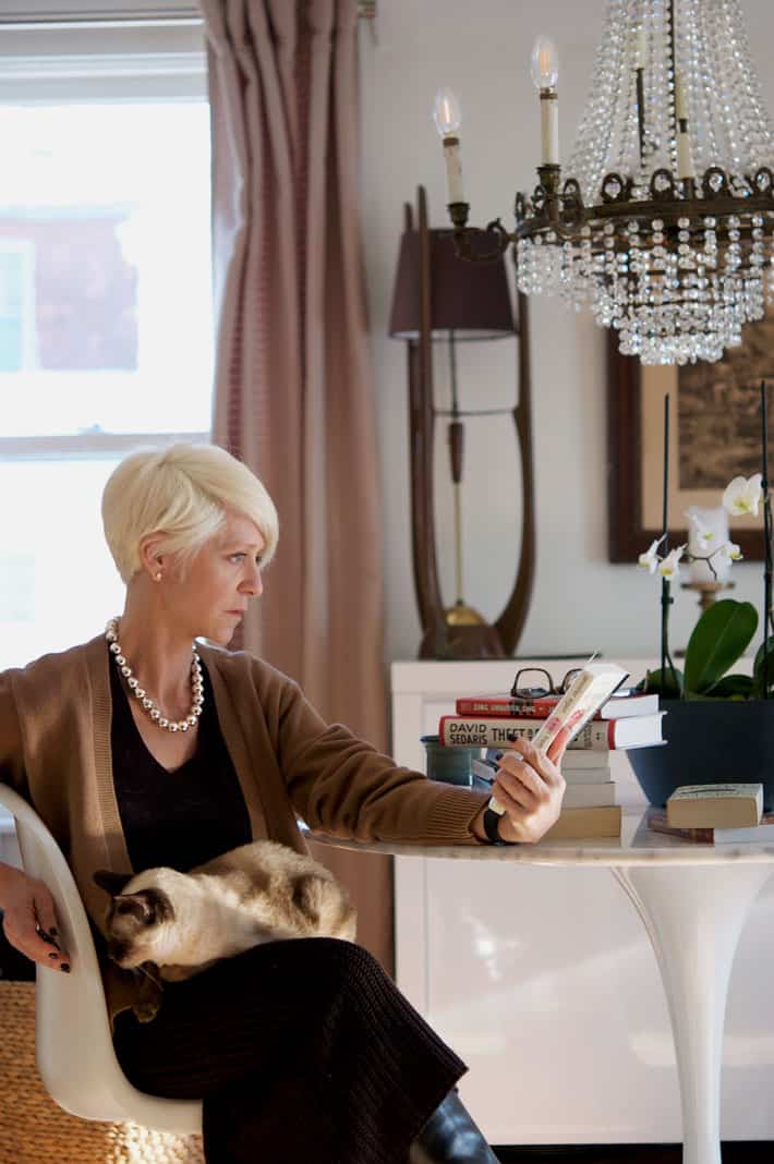 Karen Bertelsen sitting at a white marble Tulip Table with a small Siamese cat on her lap, looking at the back of a book.