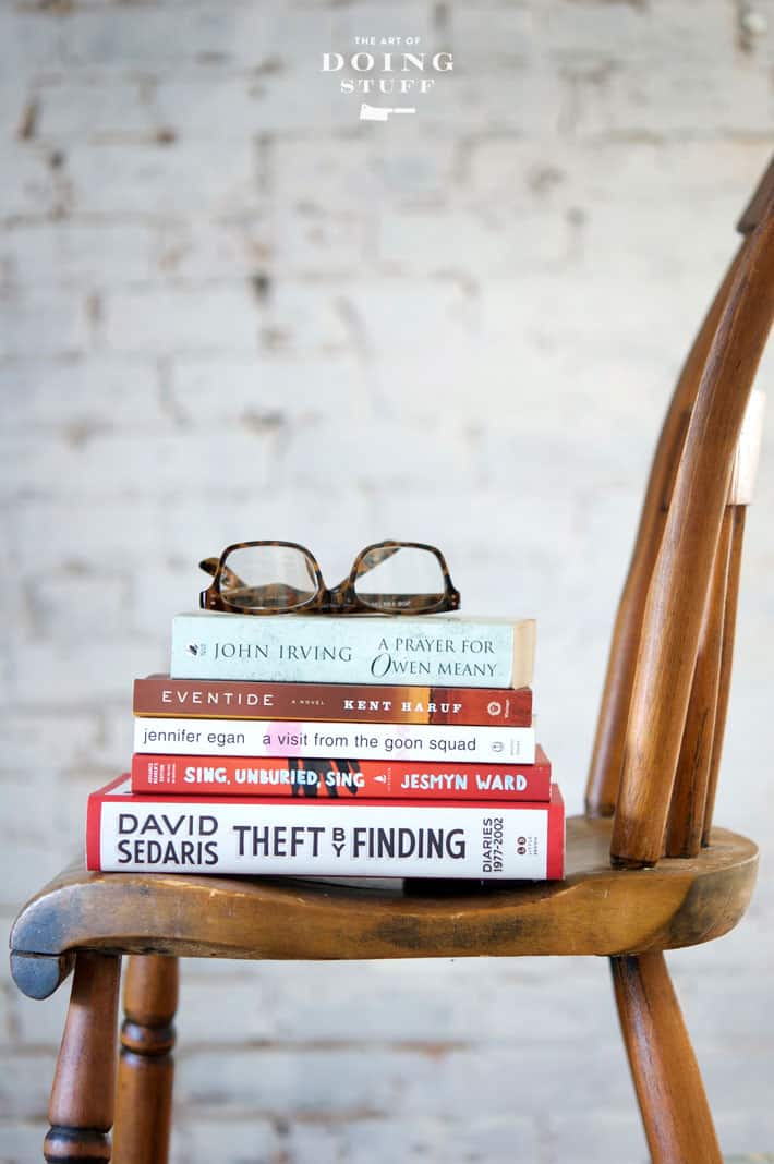 Side shot of an antique pine chair in front of a white brick wall. 5 books stacked on the chair seat topped with a pair of reading glasses.