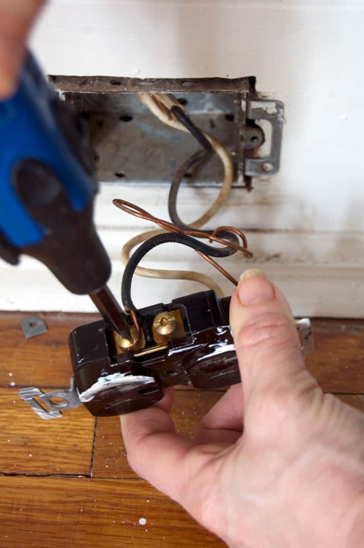Blue screwdriver unscrewing black wire from brass screws on old electrical outlet.