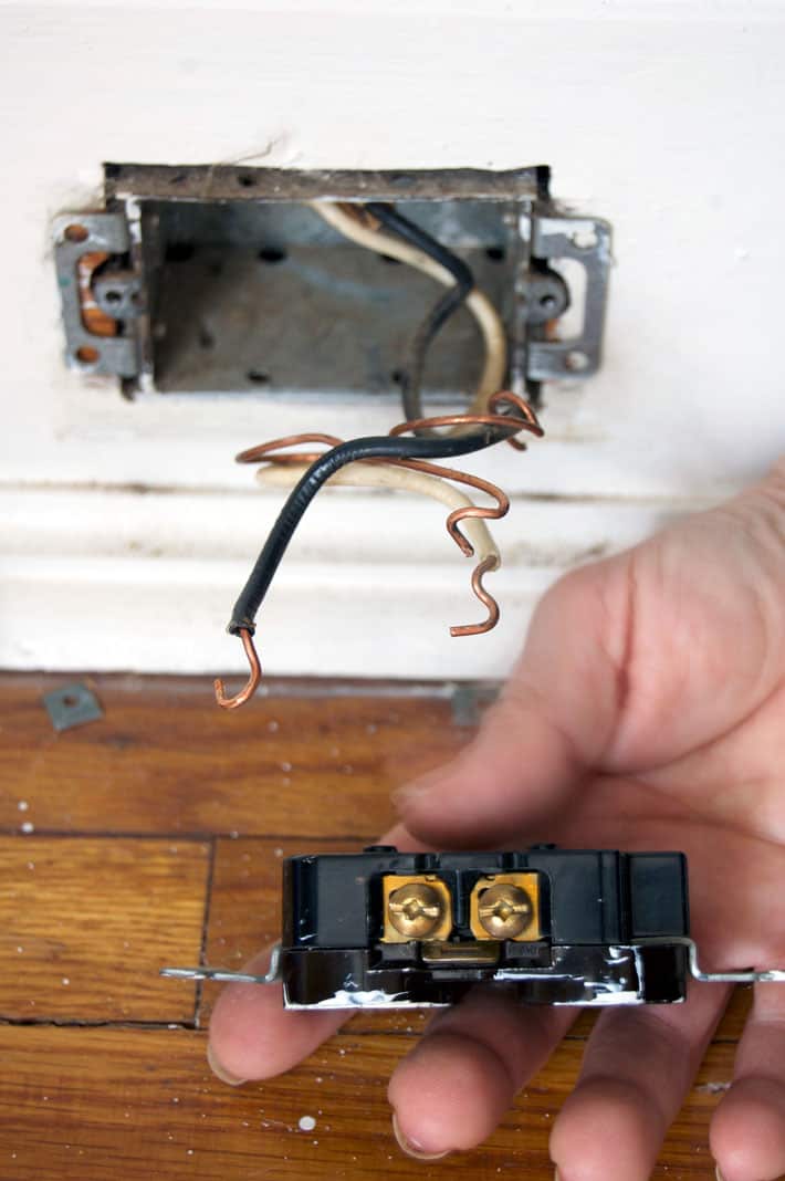 Old electrical outlet in palm of hand in front of now empty electrical box and bare wires.