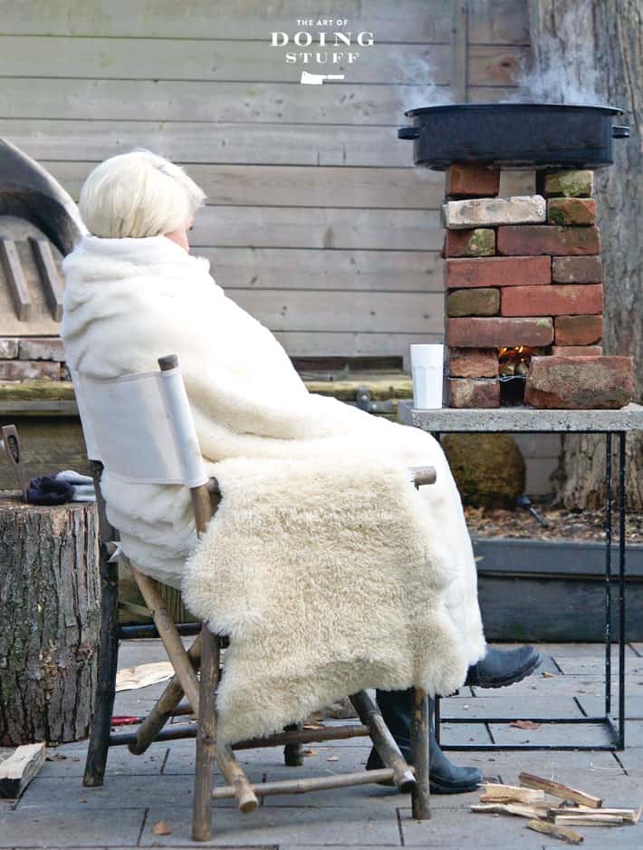 Karen Bertelsen sits outside bundled in blankets beside a brick rocket stove boiling maple syrup sap.