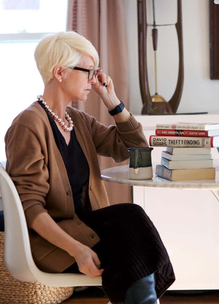 Karen Bertelsen sitting at tulip table putting on a pair of glasses, looking at a stack of books in front of her.