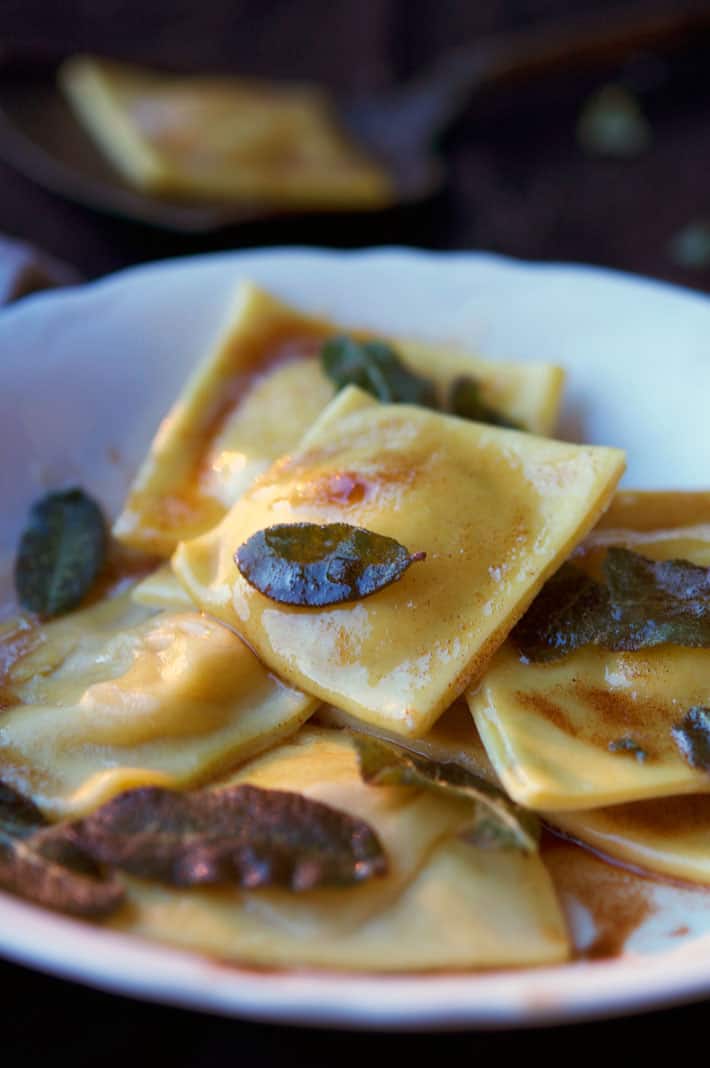 Homemade pumpkin ravioli in ironstone bowl with fried sage leaves and drizzled with browned butter.