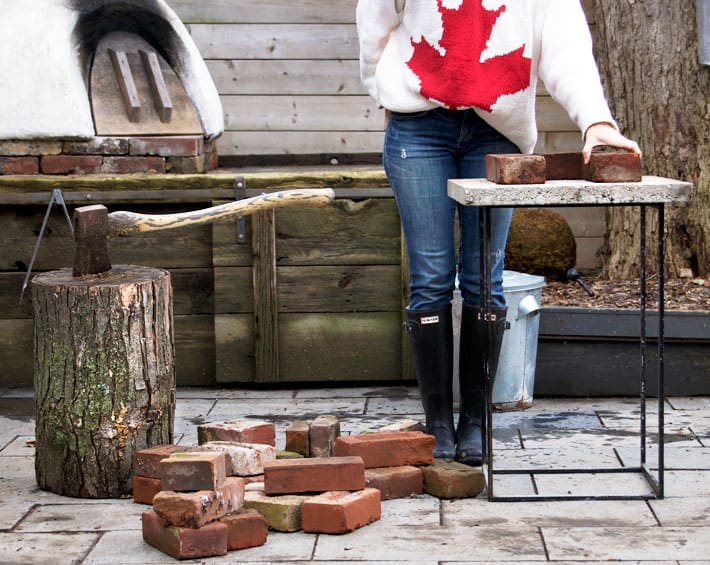 Karen Bertelsen wearing jeans and a maple leaf sweater starts assembling a rocket stove out of bricks in a backyard.