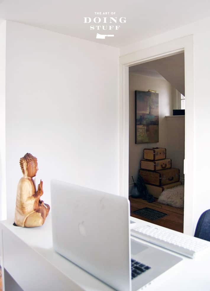 A plain white wall in a home office, with laptop and wood buddha on desk.