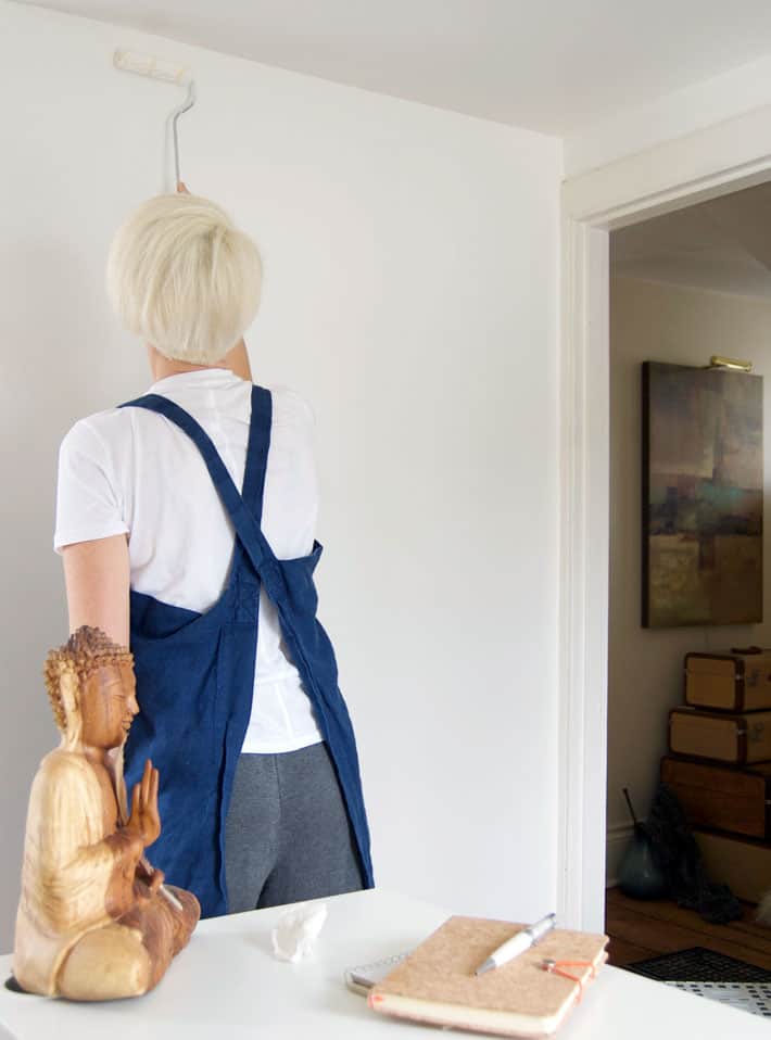 Small office wall being painted white by woman in blue pinafore.