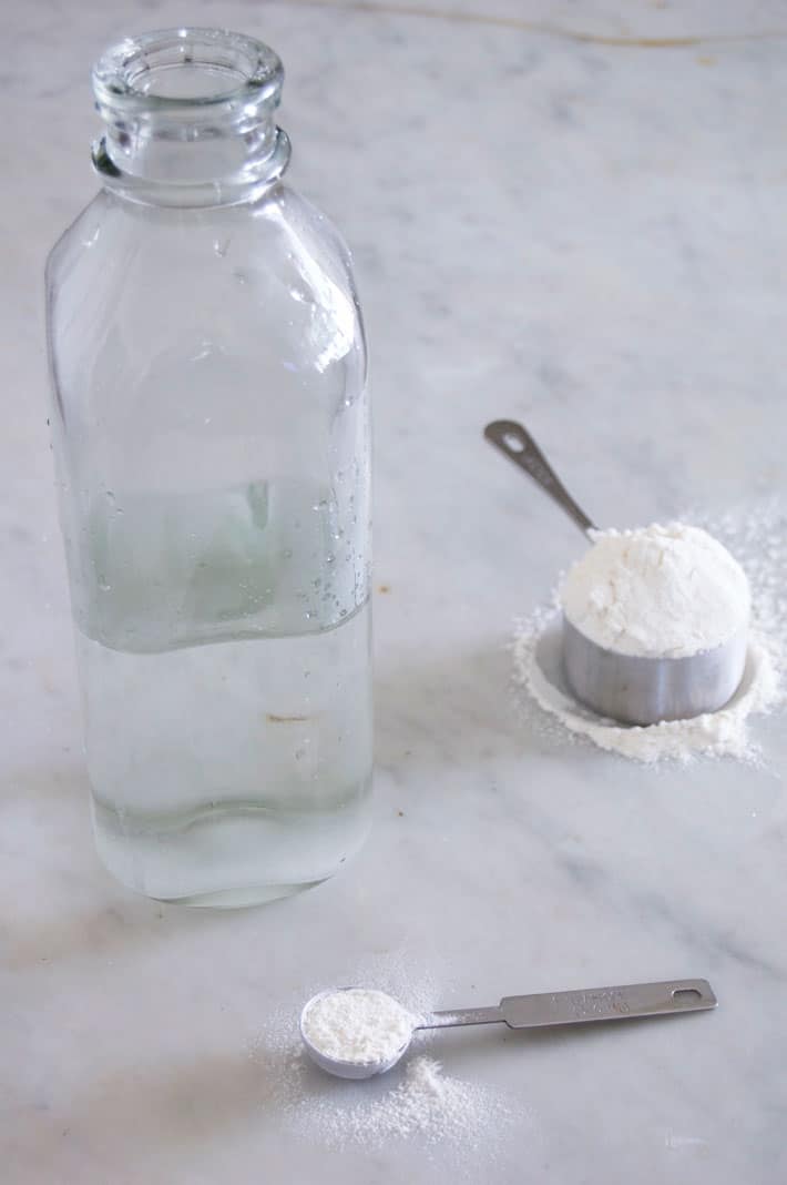 Glass milk bottle with teaspoon filled with flour beside it on marble counter.