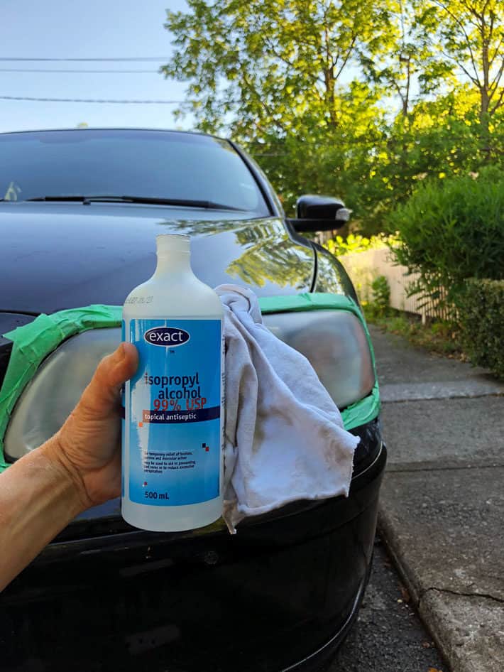 How to clean headlights like the professionals do. A bottle of isopropyl alcohol held up in front of a black car and headlight.