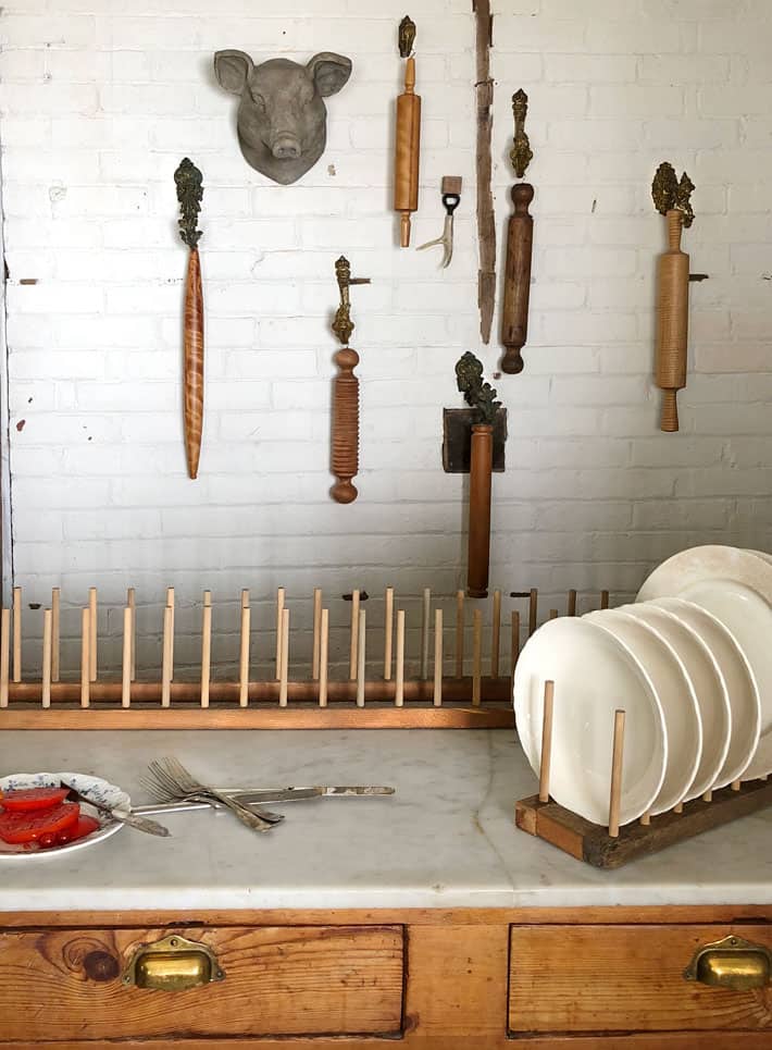 2 large plate racks on marble island top with white brick wall covered in hanging rolling pins in background.