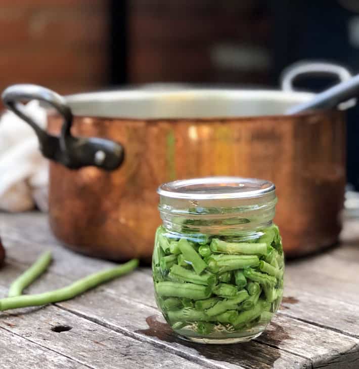 Small mason jar filled with fresh cut green beans in front of copper pot.