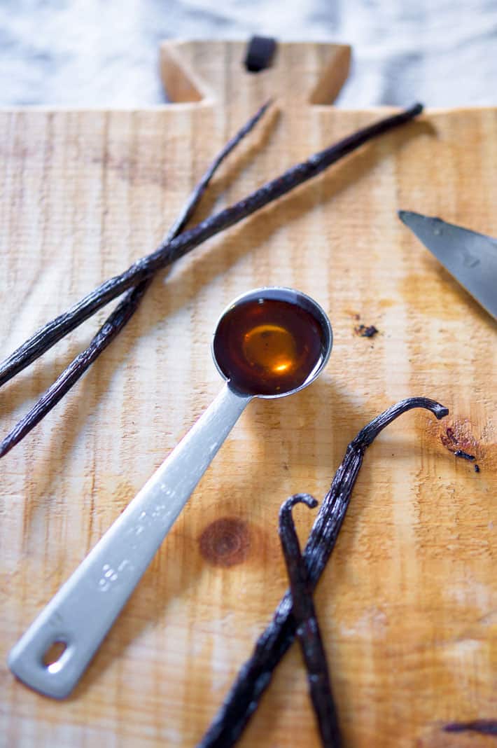 Dark homemade vanilla extract show in a measuring spoon with vanilla beans scattered around it.