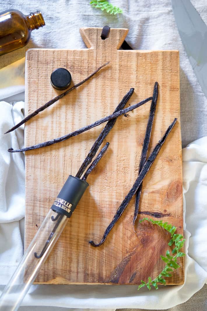 Vanilla beans in a glass tube laid out on a wood cutting board set on linen.