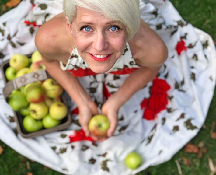 Karen Bertelsen in red rose covered vintage dress, sitting on grass with a basket of ripe apples.