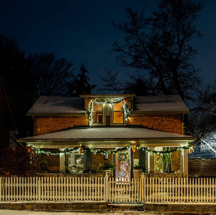 Cozy evening shot of English style brick cottage decorated with Christmas garland and white lights standing behind a white picket fence.