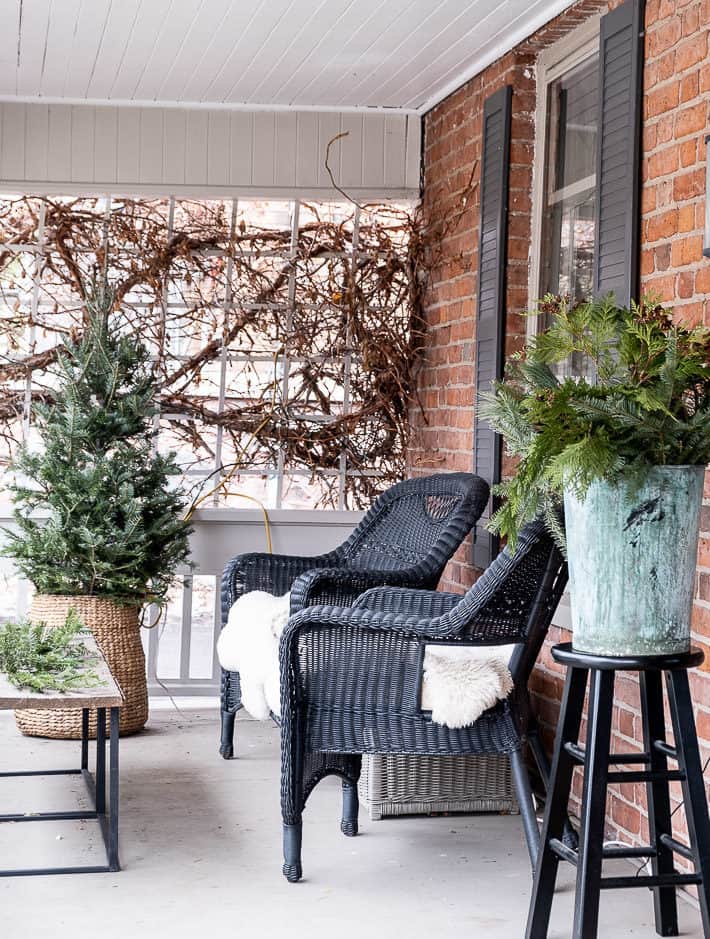 A small Christmas tree in a wicker basket set beside two black wicker chairs with sheepskin throws on a front porch.