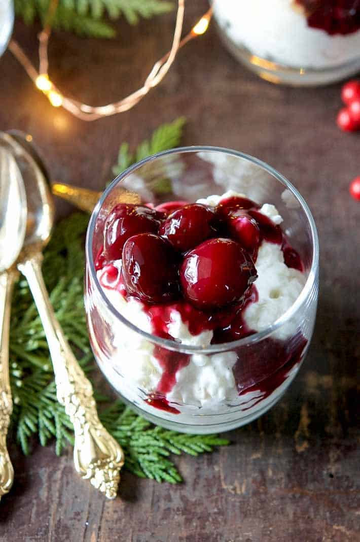 Risalamande in a clear glass topped with a bright red cherry sauce, on a wood table with cedar branches around.