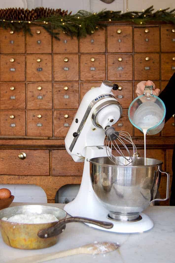 Pouring whipping cream into a stand mixer with an antique wood cabinet with garland in the background.