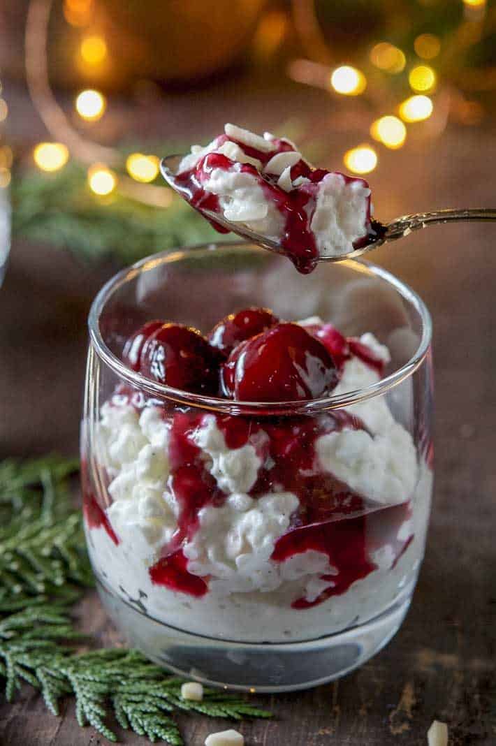 A big spoonful of rice pudding dripping with cherry sauce with glowing twinkle lights and cedar branches in the background.
