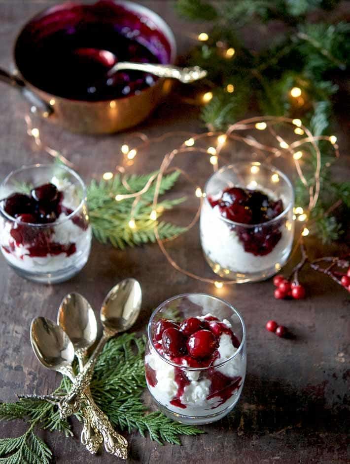Cheery glasses of danish rice pudding, Risalamande with cherry sauce on top, on a wood table surrounded with cedar branches and twinkle lights.