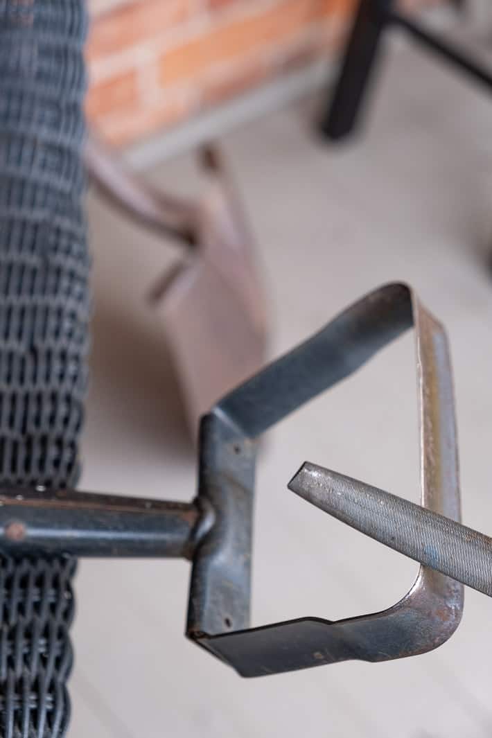 Sharpening a stirrup hoe with a metal bastard file while it rests on a black wicker chair on a front porch.