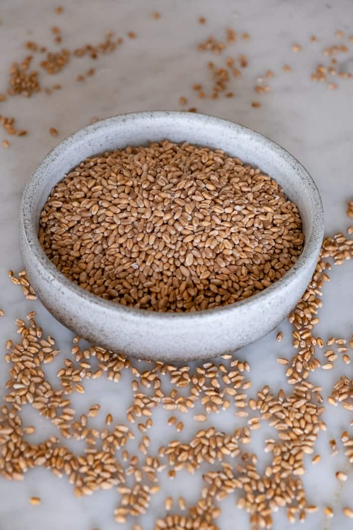 Wheat berries fill a clay bowl with some spilled over onto a marble countertop.