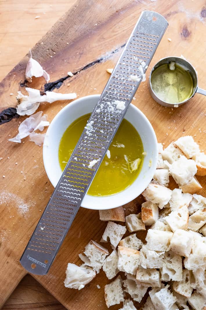 Metal rasp resting on rim of white bowl, grating fresh garlic cloves into mixture of butter and olive oil, cubed bread for croutons to the side.