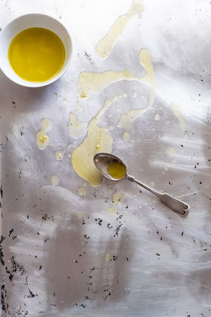 Melted butter, garlic and olive oil in a white bowl being drizzled on a well used baking sheet.