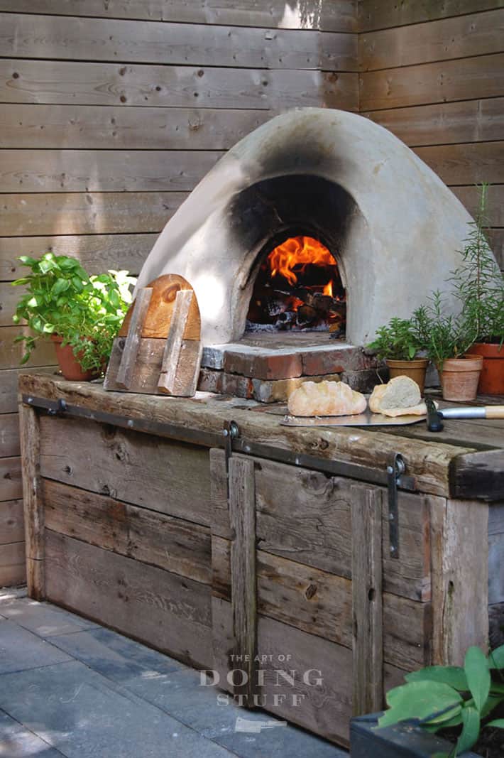 DIY cob pizza oven with a barnboard and antique brick heart, shows a roaring fire inside and freshly baked bread.