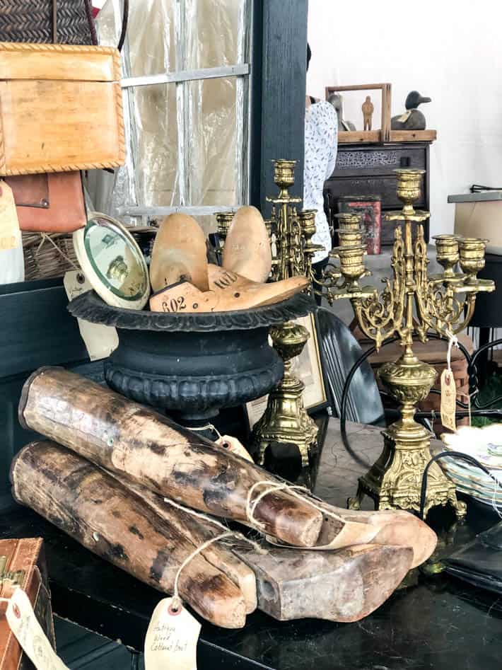 Assortment of antique show finds sitting on a table including tall boot forms, shoe forms in a black cast-iron urn and antique brass candelabras.