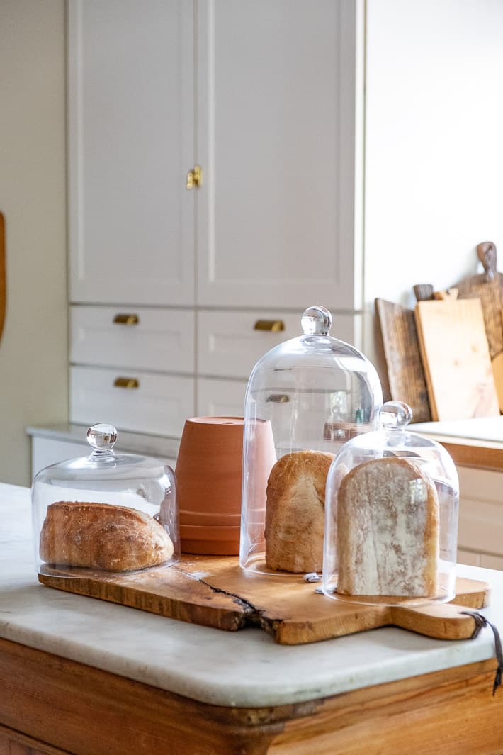 Want fresh bread? Try one of these methods. Experimenting with storing bread shows half loaves stored under glass domes and a clay pot on a white marble kitchen island.
