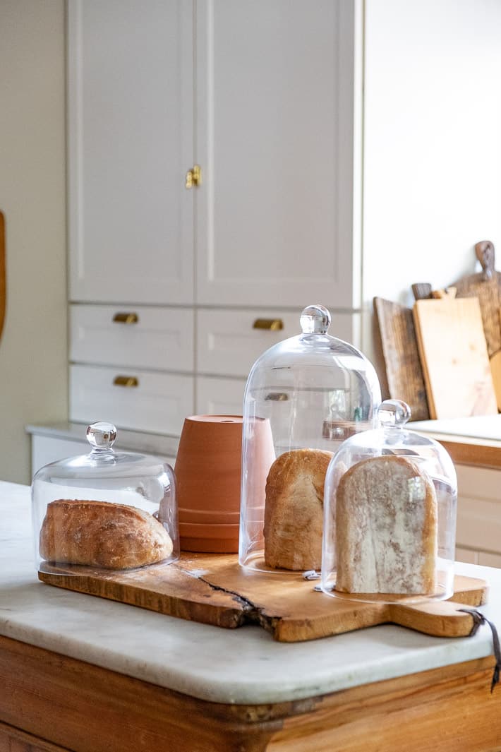 Experimenting with storing bread shows half loaves stored under glass domes and a clay pot on a white marble kitchen island.