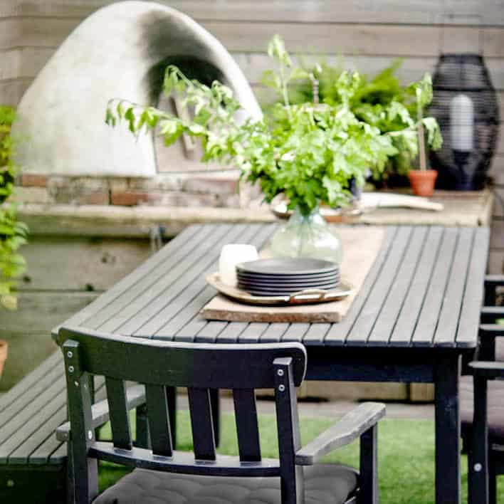 Elegant black, wood outdoor table set up in front of a homemade cob pizza oven and hearth.