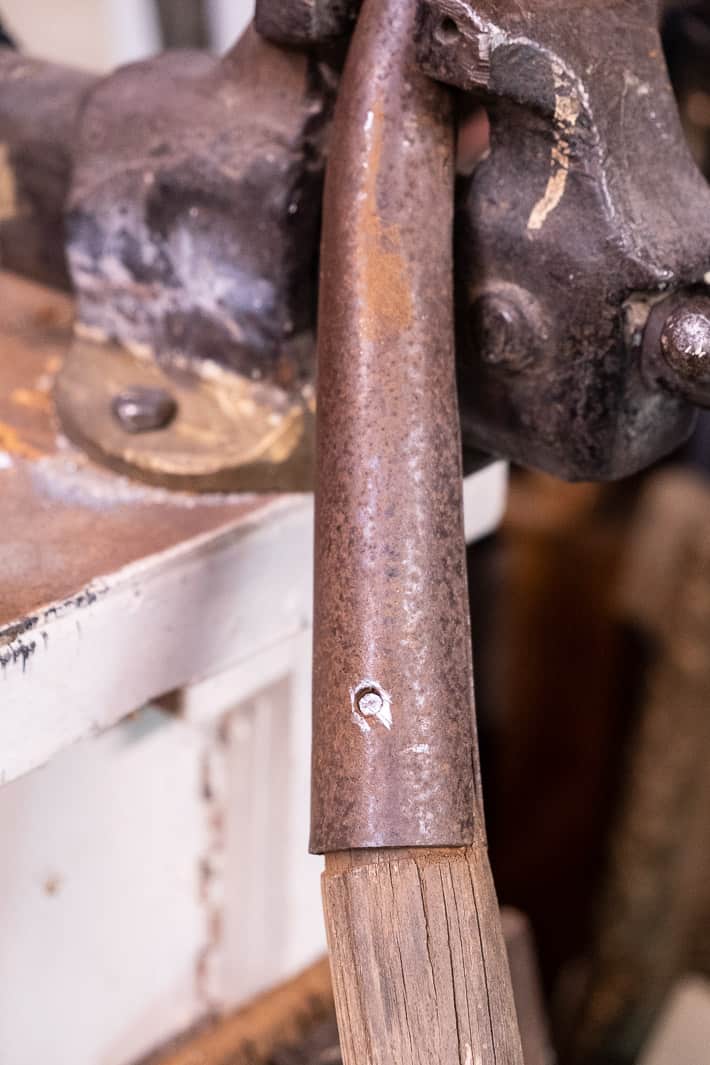 Rusted shovel handle being held tightly in a vice showing rivet with the head removed by hawcksaw.