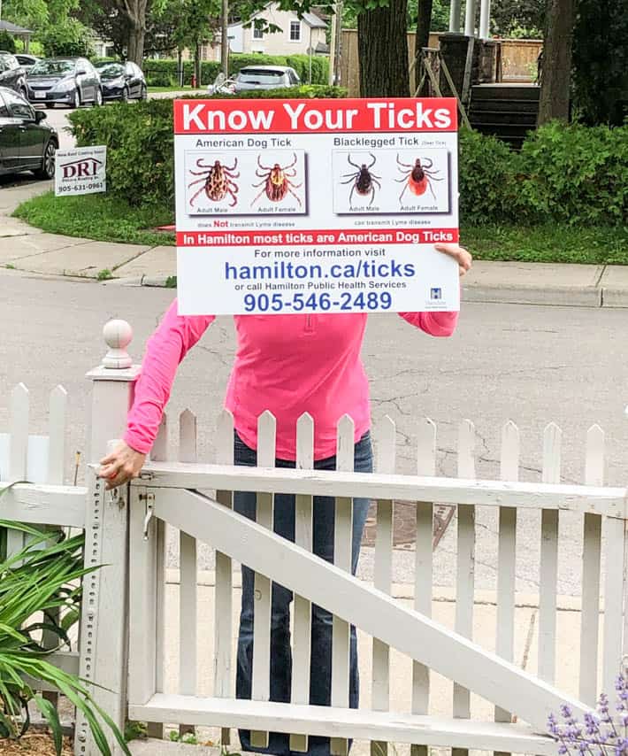 Tick identification sign being held up by a woman in a pink shirt as she enters picket fence gate.