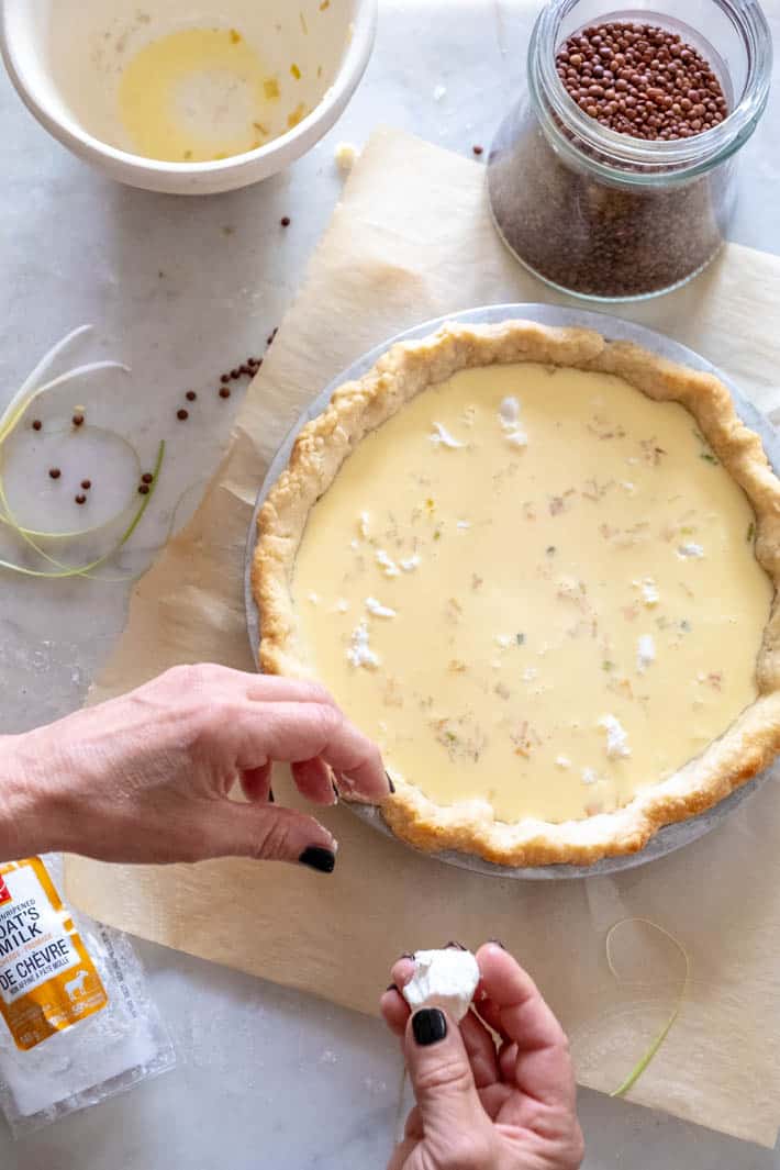 Crumbling goat cheese on top of unbaked quiche, with a jar of dried beans for use as pie weights and an ironstone bowl that held the filling.